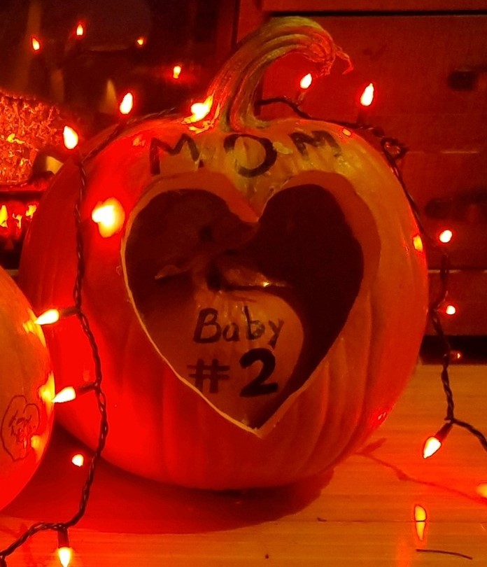 A pumpkin that says Baby number 2 is nestled inside a heart-shaped hole carved into a bigger pumpkin.